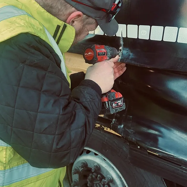 Person i refleksklær bruker drill for å feste eller reparere del på svart tilhenger ved hjul.