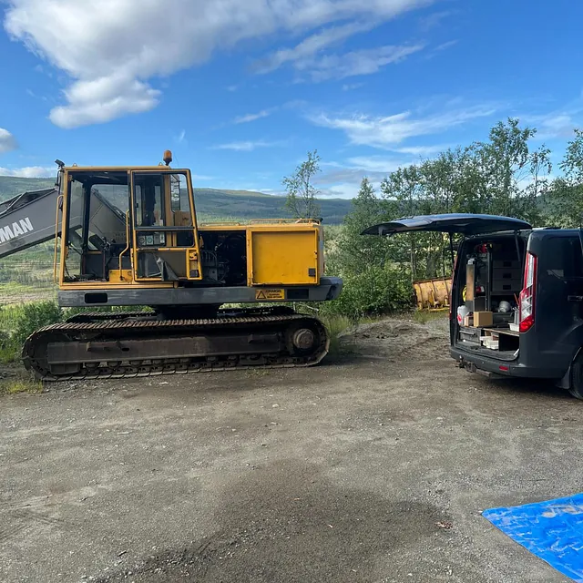 Stor beltegående gravemaskin med åpne motorluker står parkert ved servicebil på grusplass.