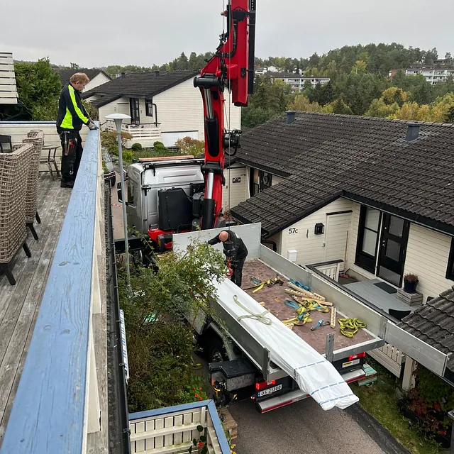 Kranbil løfter materialer opp til terrasse mens person overvåker løftet fra rekkverk.