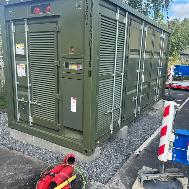 Stor grønn teknisk container står på betongfundament med røde rør og sperremateriell foran.