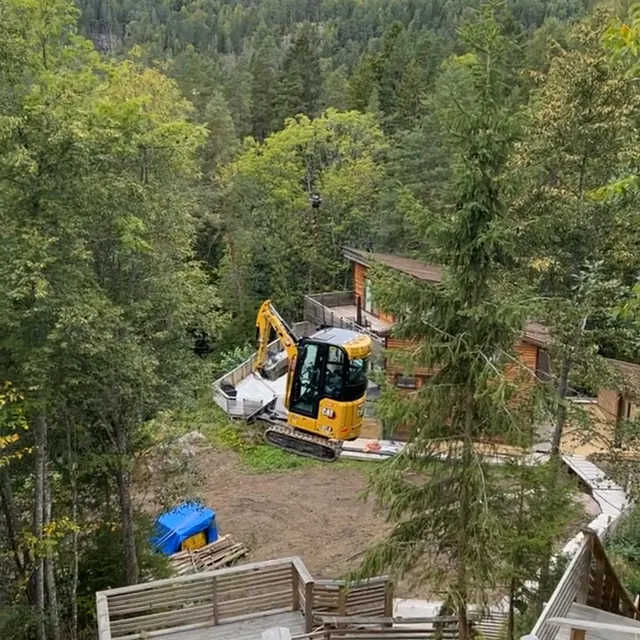 Gravemaskin står ved hytte i skogsterreng med trapper og gangveier i tre som fører ned mot bygget.