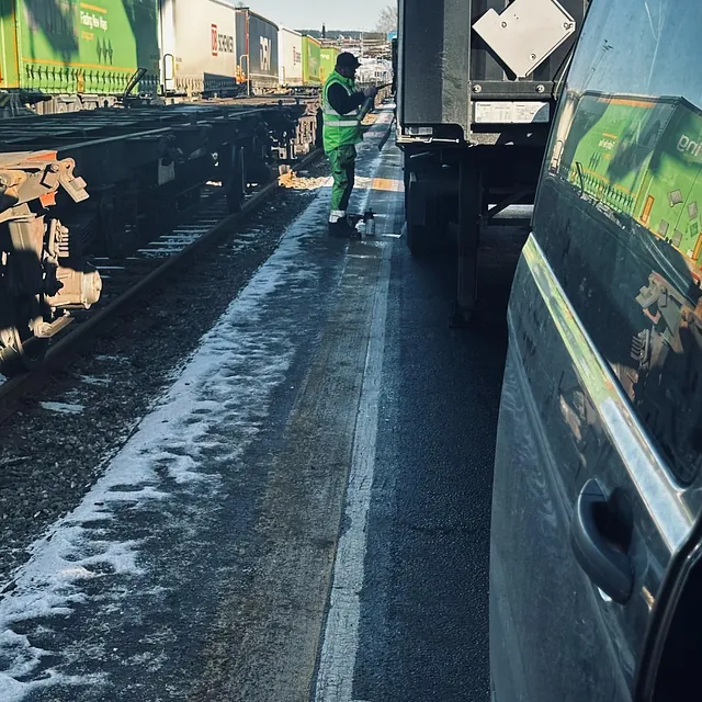Arbeider i refleksvest inspiserer godsvogner og containere ved togspor i et logistikkområde.
