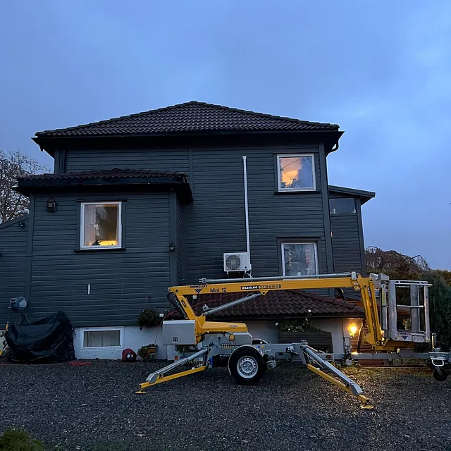 Tilhengerlift parkert foran mørkt hus i skumring, med utelys og varmepumpe på veggen.