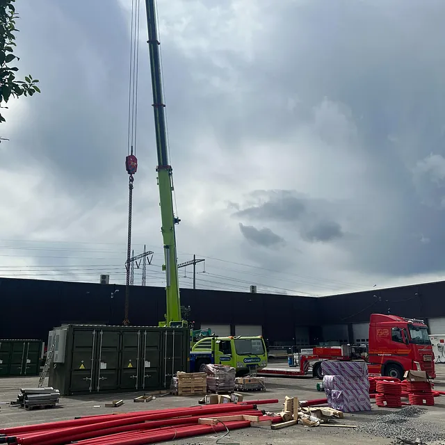 Kranbil løfter grønn teknisk container ved industriområde, med rør og byggevarer på bakken.