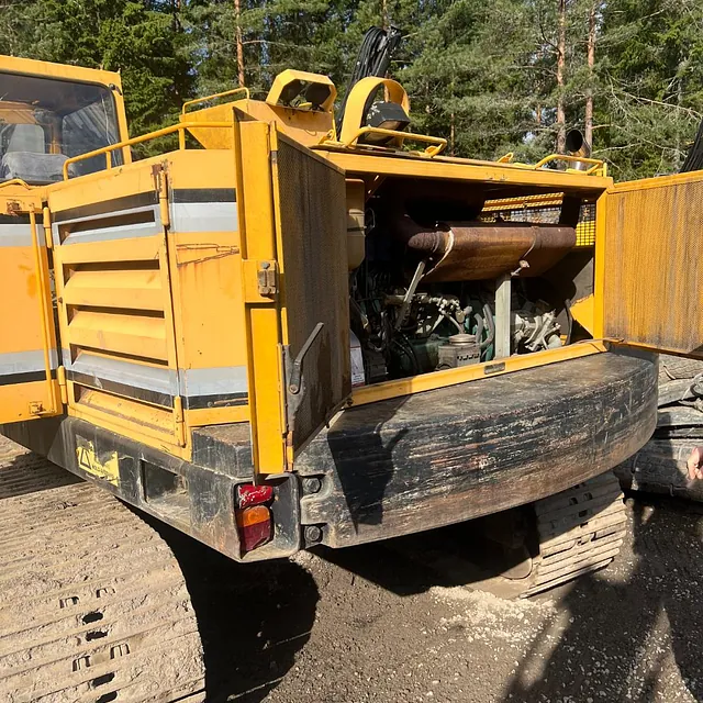 Gul anleggsmaskin med åpne motorluker står parkert på byggeplass omgitt av skog.